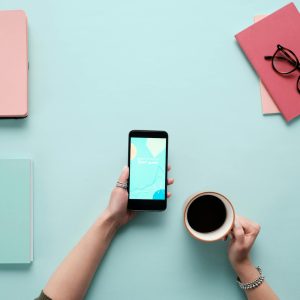Overhead view of a stylish workspace featuring a smartphone, coffee, and notebooks on a pastel background.