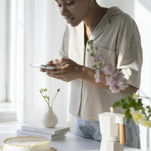 Woman capturing a stylish coffee setup in a sunlit kitchen with flowers and cake.